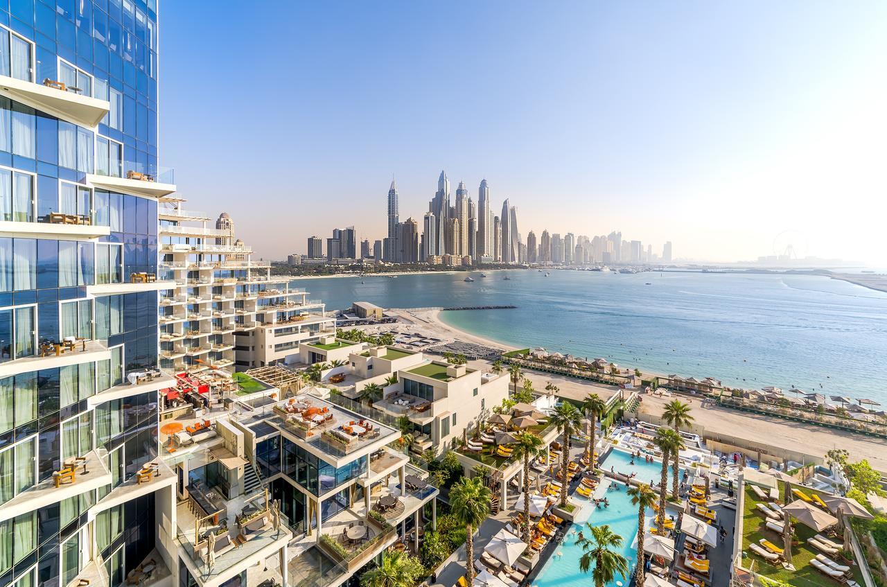 Five Palm Jumeirah Dubai Esterno foto The photo depicts a stunning seaside view featuring modern architectural structures. In the foreground, there are luxurious buildings and a pool area surrounded by palm trees. In the background, you can see a skyline of tall, contemporary skyscrapers